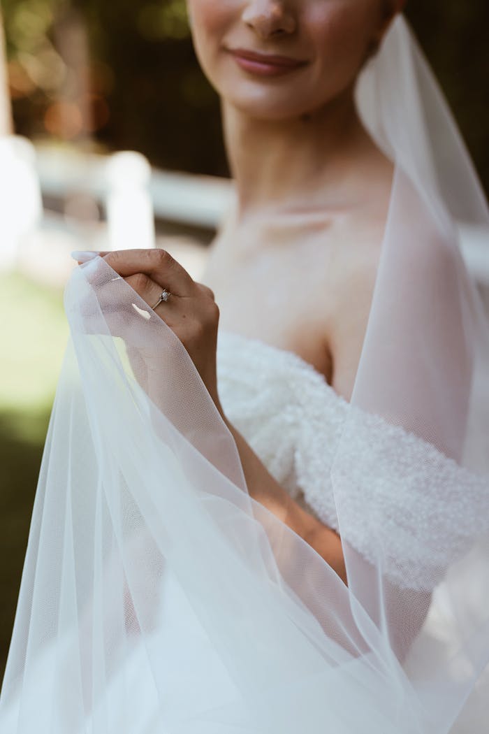 Bride in a delicate wedding gown holding her veil outdoors on a sunny day.