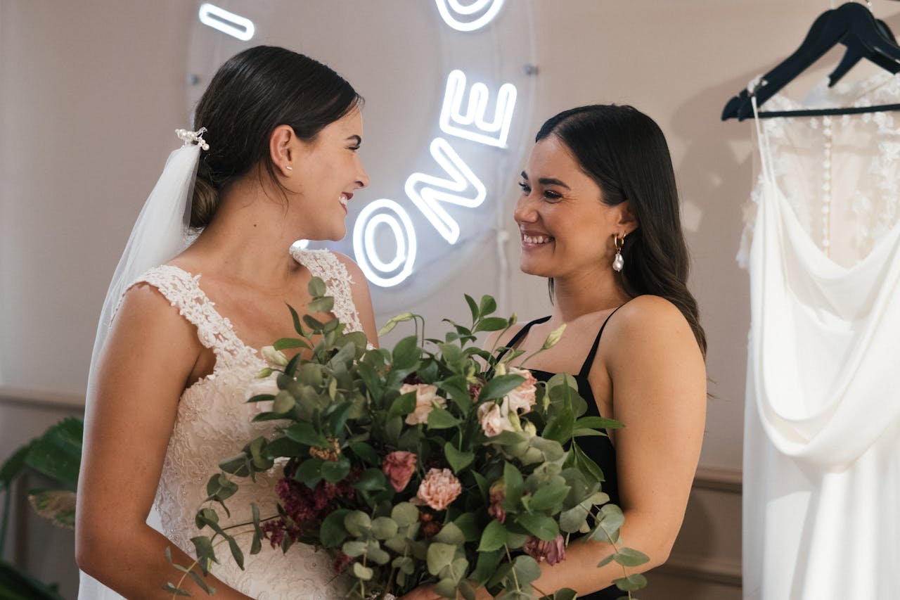Two women smiling and holding a bouquet on a wedding day indoors.